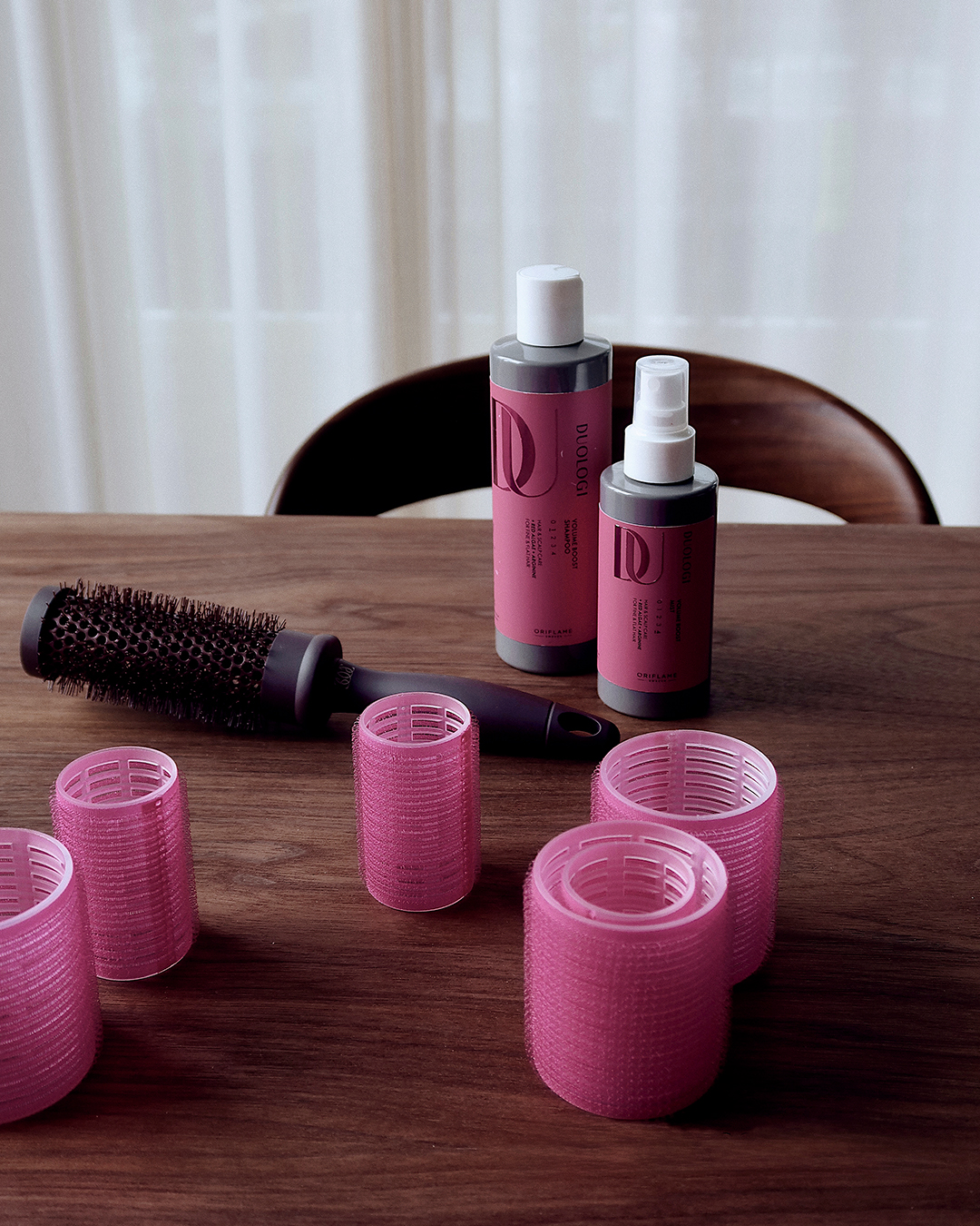 Escova redonda, rolos de cabelo e produtos Duologi Volume Boost sobre uma mesa.