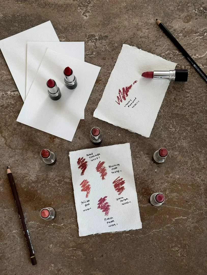Batons em vários tons de vermelho e rosa com amostras de cor sobre papel, dispostos numa superfície de pedra.