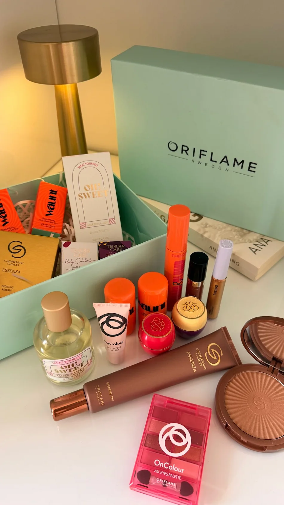 Caixa Oriflame com vários produtos de maquilhagem, fragrâncias e cuidados de beleza dispostos sobre uma mesa.